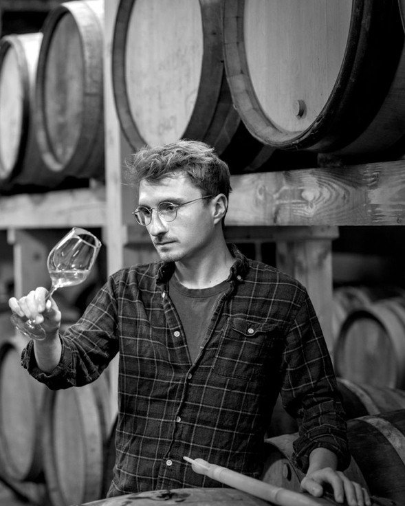 Portrait en noir et blanc d’Adrien Pestiaux examinant un verre de vin blanc devant des fûts